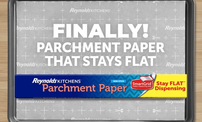 Reynolds Stay Flat Parchment Paper box sitting on top of a parchment lined baking sheet surrounded by cookies