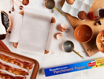 Bacon, eggs, measuring cups and other ingredients laid out on a table to show the process of baking brownies