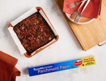 Cooked Bacon Bourbon Brownies in a parchment lined 8x8 pan alongside plates, napkins, forks and a box of Reynolds Kitchens Stay Flat parchment paper