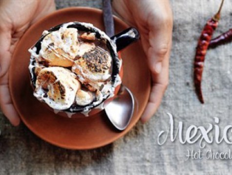 Hands holding a cup of hot chocolate topped with toasted marshmallows and spices.