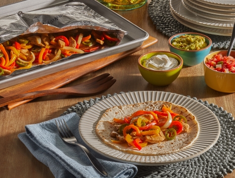 Grilled chicken fajitas sitting on a wooden table with sour cream, guacamole and salsa toppings alongside an open Reynolds grill bag with additional servings inside.