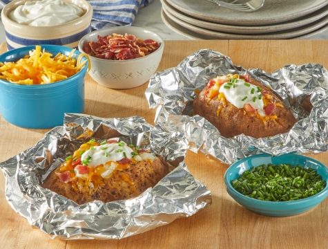 Loaded baked potato sitting in an aluminum foil sheet alongside small bowls of shredded cheese, bacon bits and sour cream