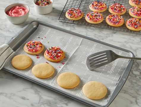 Sugar cookies with pink frosting and rainbow sprinkles sitting on a parchment lined baking sheet and wire cooling rack