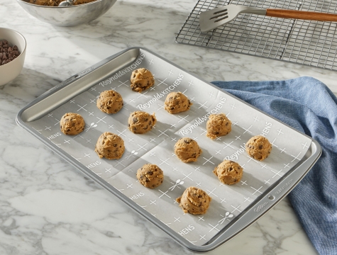 Balls of chocolate chip cookie dough sitting on a parchment lined baking sheet alongside a wire cooling rack and bowl of cookie dough