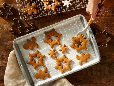 A person using a spatula to transfer holiday shaped gingerbread cookies from a parchment lined baking sheet to a wire cooling rack