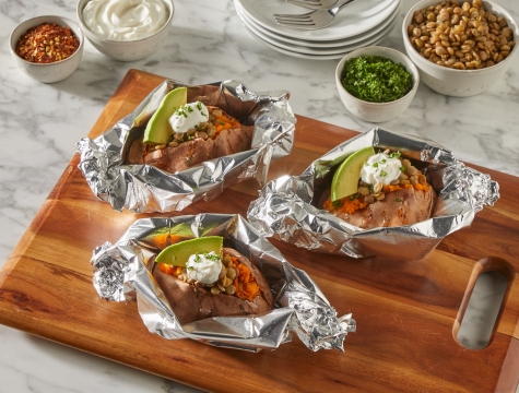 Three sweet potatoes partially wrapped in foil sitting on a kitchen counter with a variety of toppings