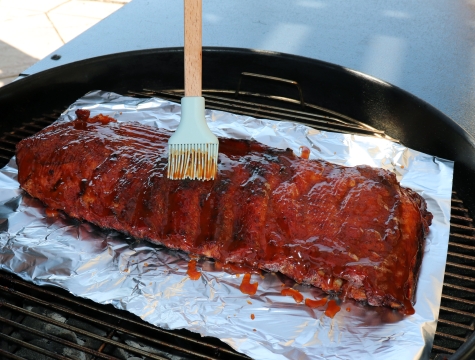 Person using a brush to add BBQ sauce to a rack of ribs sitting on a sheet of foil on a charcoal grill