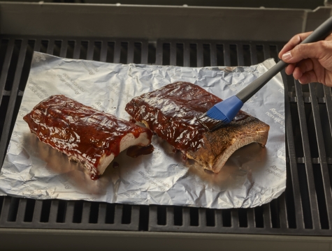 person using a silicone basting brush to coat bbq sauce on ribs sitting on a sheet of foil on the grill