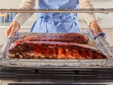 person pulling out a foil lined sheet pan from the oven topped with a rack of ribs 