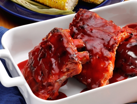 ribs sitting in a white ceramic dish alongside a plate of corn on the cob