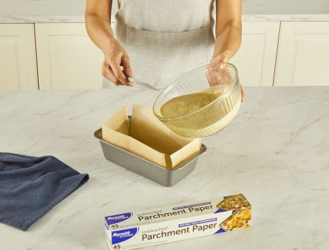 person pouring batter into a loaf pan lined with a parchment paper sling