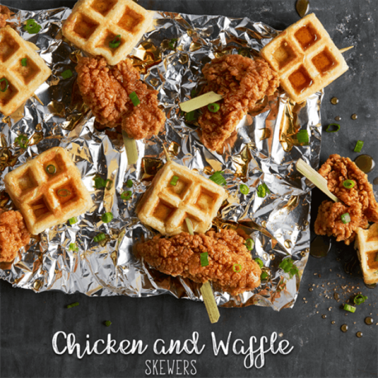 Skewers threaded with fried chicken and small square waffle sections topped with maple syrup and scallions placed on foil.