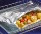 Open aluminum foil grill bag featuring corn, bell peppers, brussels sprout and potatoes sitting on a baking sheet in an open oven
