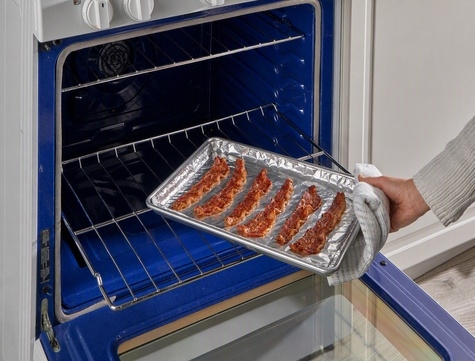 person removing a sheet pan of bacon lined with aluminum foil from the oven