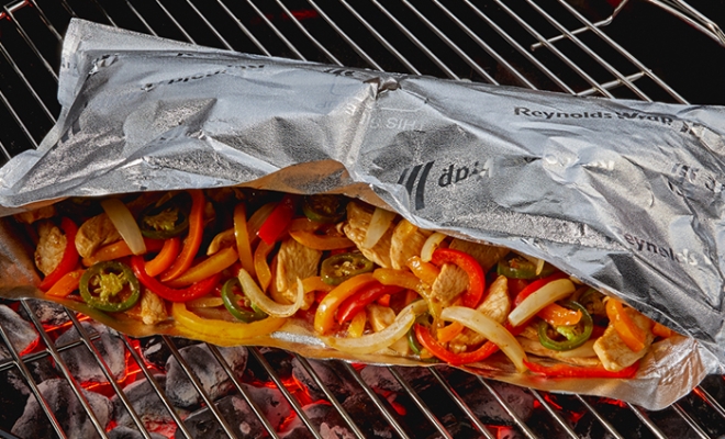 Open aluminum foil grill bag with grill chicken, onions and bell peppers inside sitting on a charcoal grill