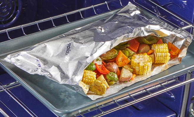 Open aluminum foil grill bag featuring corn, bell peppers, brussels sprout and potatoes sitting on a baking sheet in an open oven