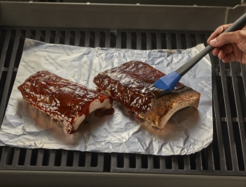 person basting a rack of ribs sitting on aluminum foil on top of grill grates