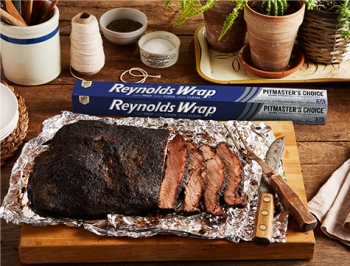 Partially sliced brisket sitting on a piece of Reynolds Wrap Pitmaster's Choice aluminum foil on a wood cutting board