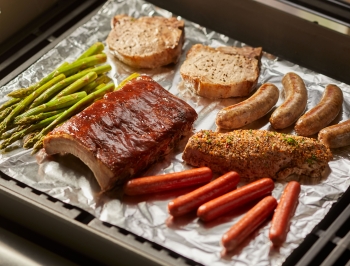 Asparagus, ribs, hot dogs, chicken and sausage sitting on a grill grate covered by aluminum foil
