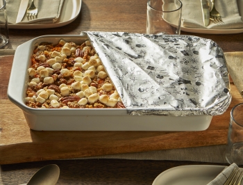 casserole dish filled with sweet potatoes topped with marshmallows sitting on a dining room table partially covered in foil
