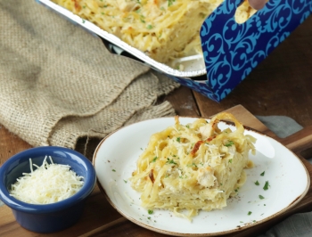 chicken alfredo fettuccini bake in a disposable aluminum container sitting on a wood table with one slice removed and sitting on a plate