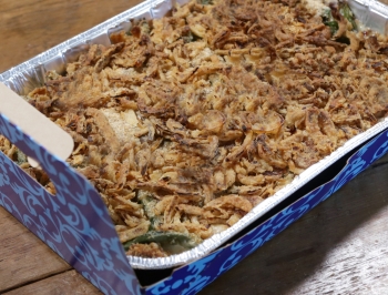 green bean casserole in a disposable pan with a carrier sitting on a wood table
