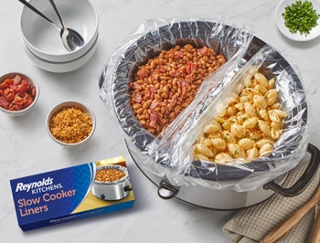slow cooker with an aluminum foil divider and both sides of the bowl lined with a slow cooker liner one side filled with mac and cheese and the other filled with baked beans