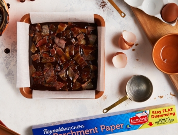 Brownie batter poured in a parchment lined 8x8 pan and a box of Reynolds Kitchens Stay Flat parchment paper alongside