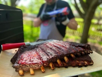 Juicy ribs resting on a wooden surface with a man holding Reynolds Wrap in the distance