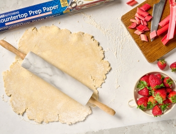 pie crust sitting on a sheet of Reynolds Kitchens Countertop Prep Paper alongside a rolling pin, strawberries, rhubarb and a cutting board